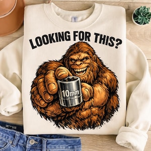 Könnte beinhalten: Cremefarbenes Sweatshirt mit dem Text "LOOKING FOR THIS?" und einer Illustration von Bigfoot, der eine 10-mm-Steckschlüssel hält. Bigfoot ist braun und orange mit einem grimmigen Grinsen.