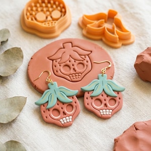 May include: A pair of dangle earrings shaped like strawberries with skull faces, featuring terracotta-colored bodies, light green leaves, and gold-toned hooks. Beside them are two orange plastic cookie cutters and a circular clay slab.