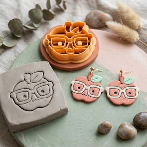 May include: An orange plastic apple-shaped cookie cutter, a gray clay slab with an embossed apple design, and a pair of peach-colored apple-shaped earrings with white glasses, a green leaf, and gold-toned posts, all arranged on a round green and pink base.