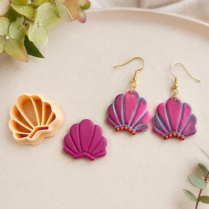 May include: A pair of dangle earrings shaped like seashells in shades of magenta and blue with small orange dots, a matching solid magenta seashell cutout, and a beige plastic seashell-shaped cutter with a ridged interior design.
