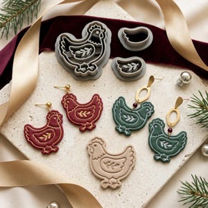 May include: A set of two grey plastic chicken-shaped cookie cutters, two pairs of dangling chicken earrings in deep red and dark green with gold-tone hardware, and one beige chicken-shaped clay piece arranged on a textured stone surface.
