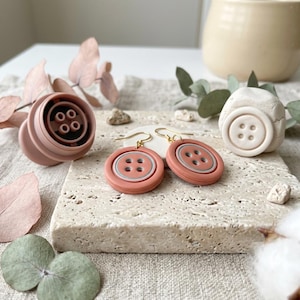 Op de afbeelding: Een paar terracotta-kleurige knoop-oorbellen met goudkleurige haakjes, voorzien van een grijze cirkel in het midden en vier gaatjes. Daarnaast liggen een bijpassende ronde vorm in terracotta en een wit kleistuk met een knoop-afdruk.