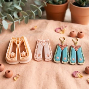 May include: A peach-colored plastic pants-shaped cookie cutter, a matching peach-colored clay pants cutout, and a pair of dangling earrings shaped like pants with teal legs, peach waistbands, and small star details on gold-toned metal hooks.