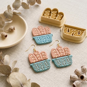 May include: A pair of dangle earrings shaped like a blue woven basket filled with three tan baguettes, accompanied by two matching plastic cutters in the same design. The earrings feature gold-toned metal hooks for attachment to the ears.