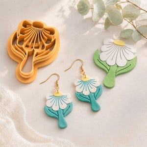 Puede incluir: Un cortador de galletas amarillo con forma de flor, un par de pendientes colgantes con dijes de flores blancas y verde azulado con centros amarillos en ganchos dorados, y una pieza de arcilla plana con forma de flor blanca y verde.