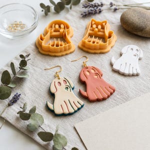 Puede incluir: Dos pendientes con forma de perro y ganchos dorados, uno en color crema y otro en terracota. Dos cortadores de galletas de plástico amarillo, una figura de arcilla blanca con forma de perro, un cuenco de cristal con anillas y lavanda.