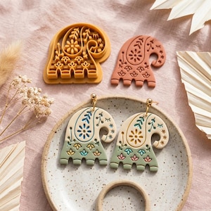 May include: A pair of cream and sage green earrings with a paisley-inspired floral pattern, accompanied by a mustard yellow plastic cutter and a terracotta-coloured clay cutout, all featuring matching intricate botanical and geometric designs.