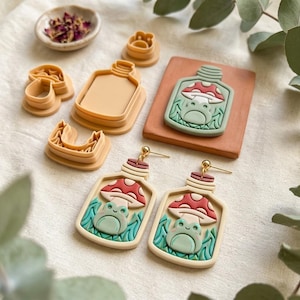 May include: A pair of dangle earrings featuring a cream-colored bottle shape with a red and white mushroom and a light green frog inside. Included are several tan plastic clay cutters in the shapes of a bottle, mushroom, and frog.