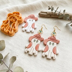Può includere: Un paio di orecchini pendenti a forma di fantasma con cappello abbinato, nei colori bianco e terracotta, appesi a ganci dorati con piccole perline turchesi. È incluso anche un tagliabiscotti arancione a forma di fantasma.