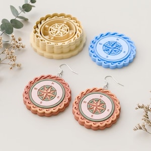 Peut inclure: Une paire de boucles d'oreilles rondes avec un motif de rose des vents et un bord festonné dans des tons rose, vert sauge et terre cuite. À côté se trouvent deux emporte-pièces circulaires, un jaune clair et un bleu clair, arborant un motif de rose des vents avec des bords festonnés.