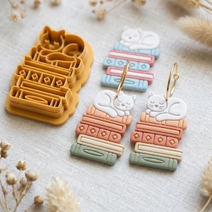 Puede incluir: Un cortador de galletas de plástico color amarillo mostaza con forma de gato durmiendo sobre una pila de libros, junto a dos pendientes a juego que muestran un gato blanco sobre una pila de libros en tonos melocotón, crema y verde salvia con aros dorados.