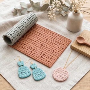 May include: A grey cylindrical texture roller with a floral pattern, a rectangular terracotta-coloured clay slab with a matching floral imprint, two light blue drop earrings with floral textures, and a pale pink circular pendant on a gold chain.