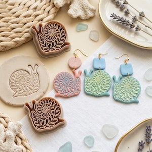 Könnte beinhalten: Zwei beige schneckenförmige Ausstechformen aus Kunststoff, eine runde Tontonne mit eingeprägtem Schneckenmotiv und drei hängende Ohrringe mit Schneckenanhängern in Rosa, Hellblau und Mintgrün mit goldfarbenen Metallhaken.