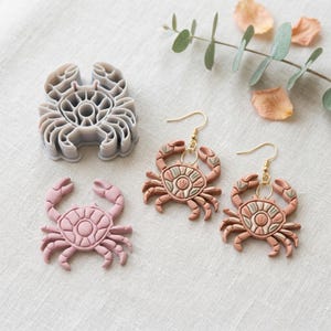 May include: A selection of crab-themed items. Includes a grey plastic crab-shaped cookie cutter, a pink crab-shaped pendant, and two crab earrings with gold-coloured hooks. The earrings and pendant are pink, tan, and green.