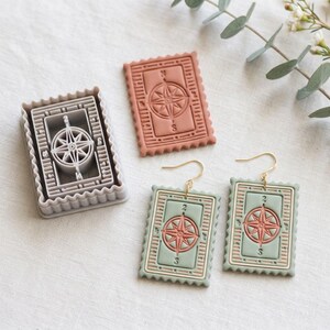 Peut inclure: Un ensemble de bijoux sur le thème du voyage et un emporte-pièce. Les boucles d'oreilles et l'emporte-pièce présentent un motif de rose des vents dans un cadre rectangulaire. Les boucles d'oreilles ont des crochets dorés. L'emporte-pièce est gris.