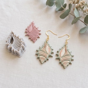 May include: A set of seashell-shaped earrings with gold-colored hooks, alongside a seashell-shaped clay cutter and two seashell-shaped clay pieces. The earrings and clay pieces feature a color palette of green, pink, and beige.