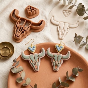 May include: A pair of dangle earrings featuring sage green bull skull pendants with light blue heart-shaped accents, paired with two brown plastic clay cutters in the shape of a bull skull and a small heart cluster for crafting jewelry.