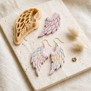 May include: A pair of wing-shaped earrings in pastel pink, blue, and yellow, a single matching wing-shaped clay piece, and a beige plastic wing-shaped cookie cutter. The earrings feature gold-toned metal hooks and detailed feather textures.