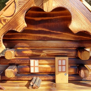 May include: A handcrafted wooden birdhouse with a rustic cabin design. The birdhouse features a sloped roof, a decorative gear-shaped element, and a small door and window. The wood has a warm, natural finish with darker accents, giving it a charming, inviting look.