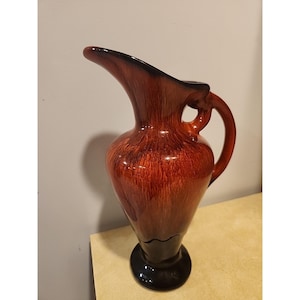 May include: A tall, glossy ceramic pitcher with a dark brown base and handle, transitioning to a rich, reddish-brown body. The pitcher has a curved spout and a rounded handle, showcasing a vintage aesthetic.