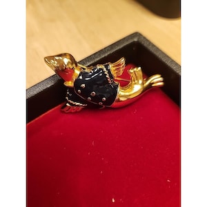 May include: A gold-toned dachshund brooch with a navy blue jacket featuring white dots. The dachshund has small wings and is lying down on a red velvet surface. The brooch is set in a black box.