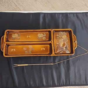 May include: A brown ceramic serving tray with three compartments and a serving fork. The tray features floral and butterfly designs in gold. The serving fork is attached to the tray with a chain.