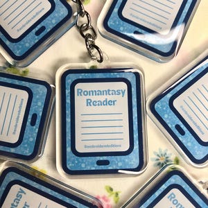 May include: A collection of rectangular acrylic keychains featuring a blue e-reader design. Each keychain displays the text Romantasy Reader, a set of horizontal lines, and the handle @embroiderededitions in blue on a light blue background.