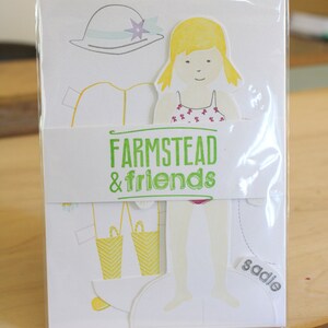 May include: A paper doll set in a clear plastic sleeve. The set includes a paper doll with yellow hair and a swimsuit, along with clothing and accessories. The words "FARMSTEAD & friends" are printed on a white band.