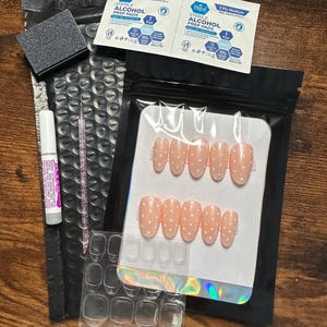 Può includere: Un kit per unghie con vari componenti. Include un set di unghie finte rosa chiaro con pois bianchi, colla per unghie, una lima per unghie, salviette imbevute di alcol e linguette per l'applicazione delle unghie. Il kit è confezionato in plastica nera e trasparente.