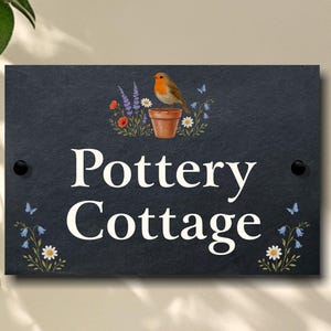 May include: A rectangular black slate sign with the words "Pottery Cottage" in white. A bird sits atop a flower pot surrounded by flowers. Butterflies and flowers decorate the corners.