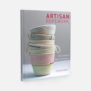 May include: A book titled "ARTISAN ROPEWORK" features a stack of handmade rope bowls in various colors. The bowls are white, pink, green, and black. The book cover also includes the text "15 3-D Stitched Rope Craft Projects" and the author's name, Jessica Geach.