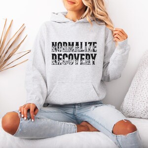 May include: A light gray hoodie with the words "NORMALIZE RECOVERY" printed in black. The hoodie has a front pocket and a hood. The person in the image is wearing blue jeans.