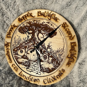 May include: A round wooden clock with a laser-cut design featuring a hobbit hole and tree. The clock face has black hands and is surrounded by text in a stylized font. The clock is on a gray, textured surface.
