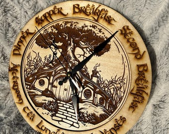 May include: A round wooden clock with a laser-cut design featuring a hobbit hole and tree. The clock face has black hands and is surrounded by text in a stylized font. The clock is on a gray, textured surface.