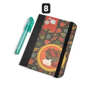 May include: A black-bound notebook featuring a dark cover with a pattern of autumn leaves and a central circular graphic of a basket of red apples, secured by a black elastic band, accompanied by a translucent teal plastic pen and the number 8.