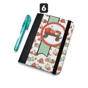 May include: A notebook with a black spine and an elastic closure, featuring a pattern of orange and green pumpkins on a white background. A central circular emblem displays a wagon filled with pumpkins. A translucent teal pen is positioned nearby.