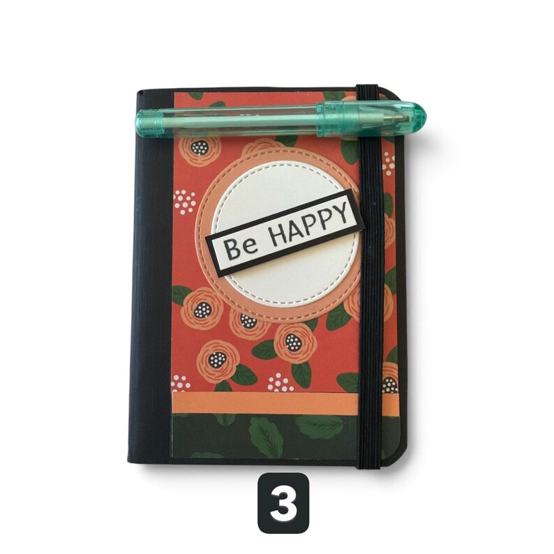 May include: A black notebook with a red floral patterned cover featuring a white circular cutout and a rectangular label with the text Be HAPPY. A translucent light blue pen is secured horizontally across the top by a black elastic band.