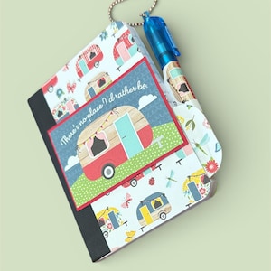 May include: A notebook and pen set with a camping theme. The notebook features a black spine and pages, with a cover adorned with colorful camper illustrations and the text "There's no place I'd rather be." A blue pen is attached.
