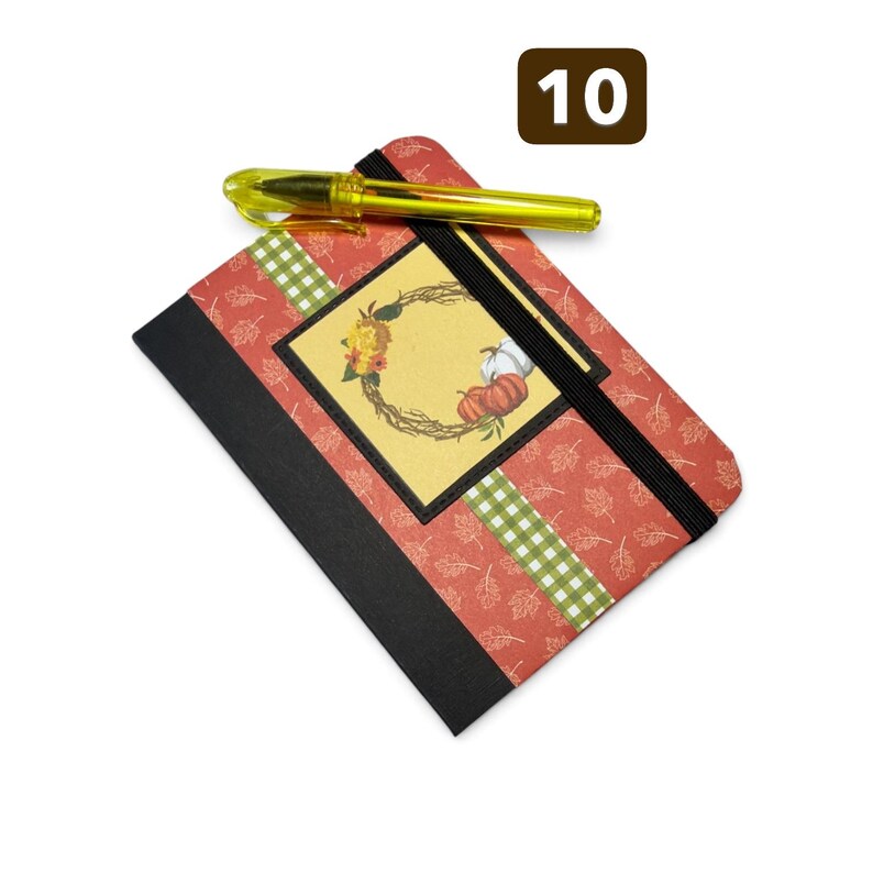 May include: A rectangular notebook with a red cover featuring a white leaf pattern, a black spine, and a green gingham stripe. A central square label displays a wreath with pumpkins and flowers. A yellow translucent pen rests on top.
