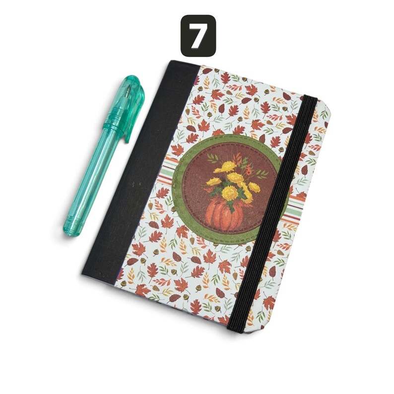 May include: A notebook with a white cover featuring a pattern of autumn leaves and acorns, a black spine, and a black elastic closure band. A circular patch displays a pumpkin and yellow flowers. A translucent teal pen is positioned next to it.