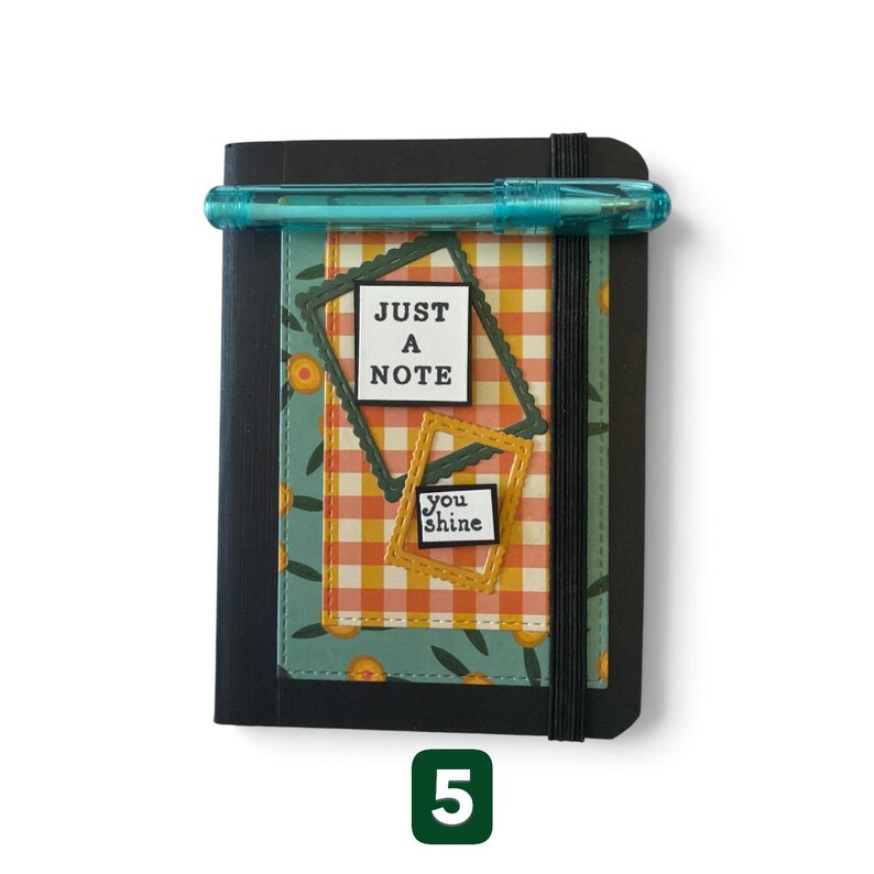 May include: A black notebook with an elastic band closure, featuring a teal floral cover insert with an orange and white gingham rectangle. Two decorative frames contain the text "JUST A NOTE" and "you shine". A translucent teal pen rests on top.