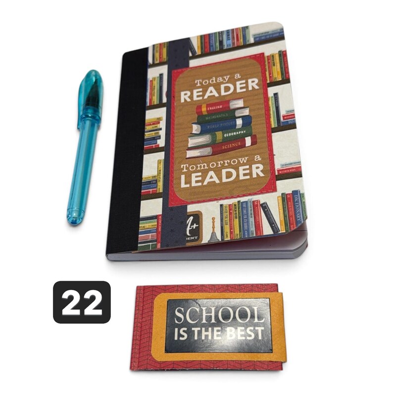 May include: A notebook with a bookshelf graphic and the text Today a READER Tomorrow a LEADER, a blue pen, and a small rectangular card with the text SCHOOL IS THE BEST and the number 22 in a black box, all displayed on a plain white background.