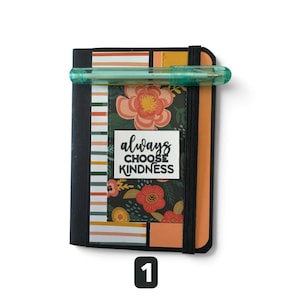 May include: A rectangular notebook with a black spine and elastic band, featuring a floral pattern in shades of orange, red, and green. A white rectangular label on the cover displays the black text "always CHOOSE KINDNESS." A translucent teal pen rests horizontally across the top.