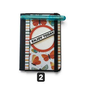 May include: A rectangular notebook with a black spine and elastic band, featuring a white cover with orange, yellow, and teal butterfly illustrations, vertical stripes, and a circular orange-bordered label with the text ENJOY TODAY, plus a teal pen.