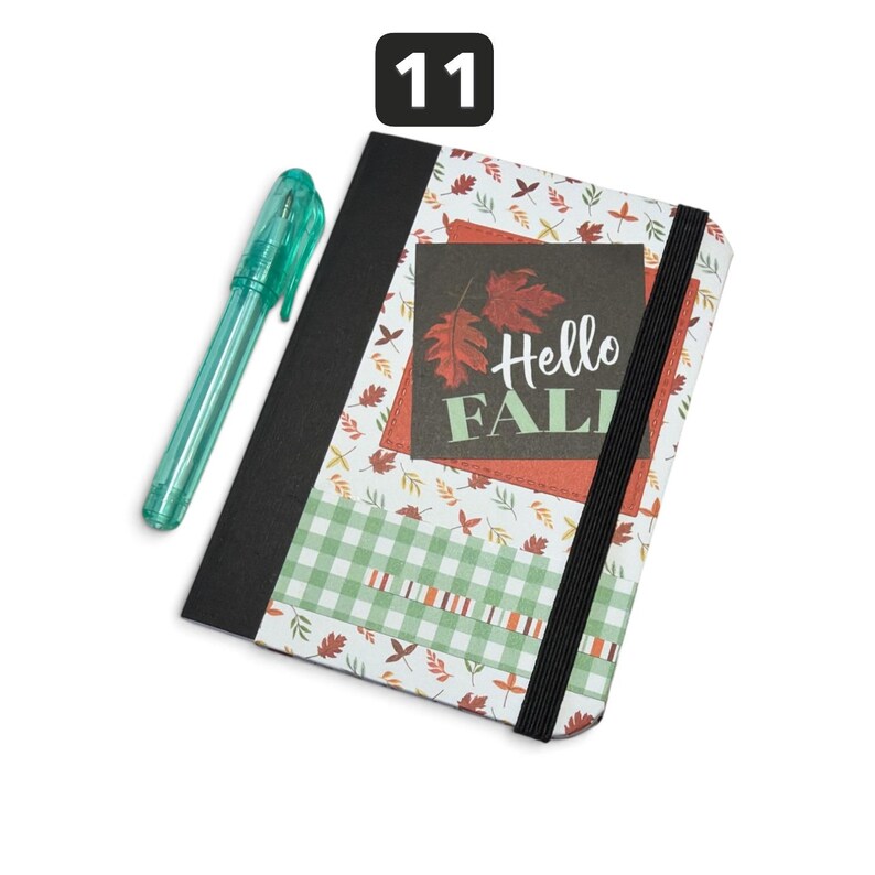 May include: A rectangular notebook with a black spine and an elastic closure band, featuring a pattern of autumn leaves and a green gingham strip. A dark square patch displays the text Hello FALL with red leaf illustrations. A teal pen is included.