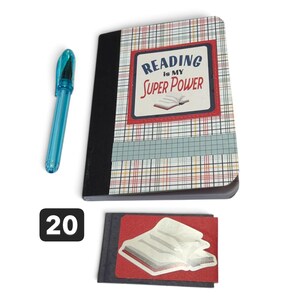 May include: A notebook with a plaid cover featuring the text READING IS MY SUPER POWER and an illustration of an open book, accompanied by a translucent blue pen and a small rectangular card displaying a graphic of an open book on a red background.