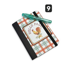 May include: A rectangular notebook with a white, orange, and green plaid cover featuring a central graphic of a rooster and the text "HELLO FALL." A black elastic band secures the notebook, and a translucent teal pen rests horizontally on top.