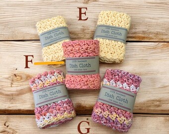 May include: Five handmade dish cloths in various colors, including pink, yellow, and purple, are displayed on a wooden surface. Each cloth is wrapped with a label that reads "100% Handmade Dish Cloth 100% Cotton Yarn."