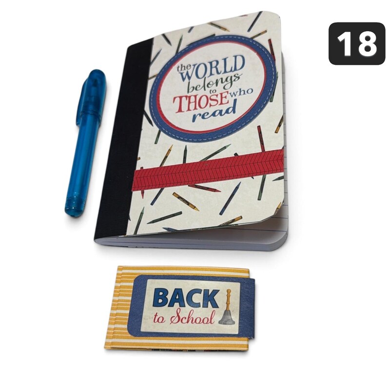 May include: A notebook with a cream cover featuring a pattern of colored pencils, a red horizontal stripe, and the text "the WORLD belongs to THOSE who read" in a blue circle, accompanied by a blue pen and a small yellow striped "BACK to School" card.