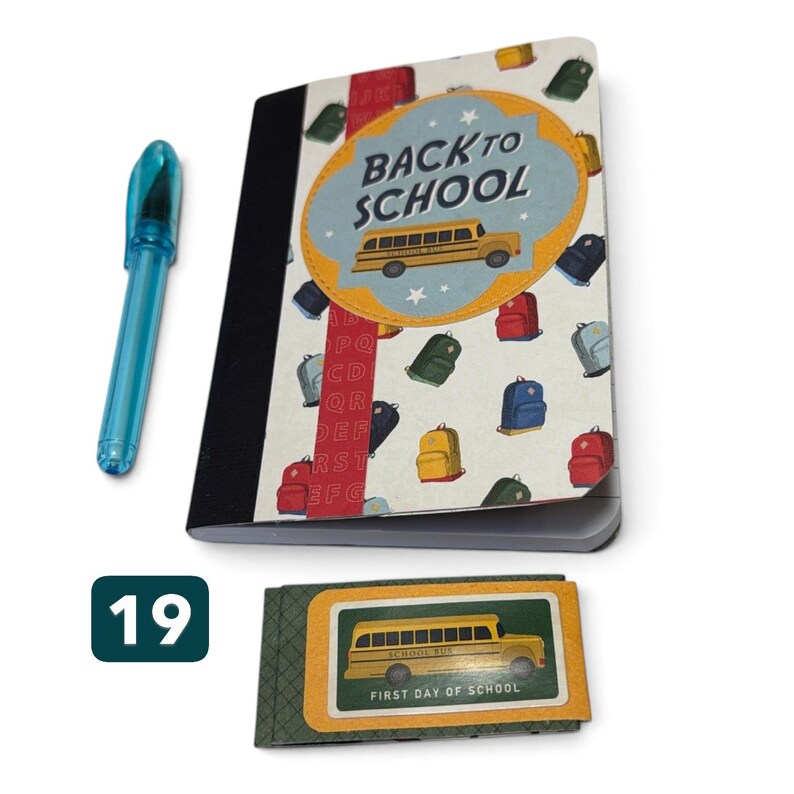May include: A notebook with a white cover featuring a yellow school bus, the text BACK TO SCHOOL, and various colorful backpacks, alongside a translucent blue pen and a small rectangular card with a yellow bus and the text FIRST DAY OF SCHOOL.